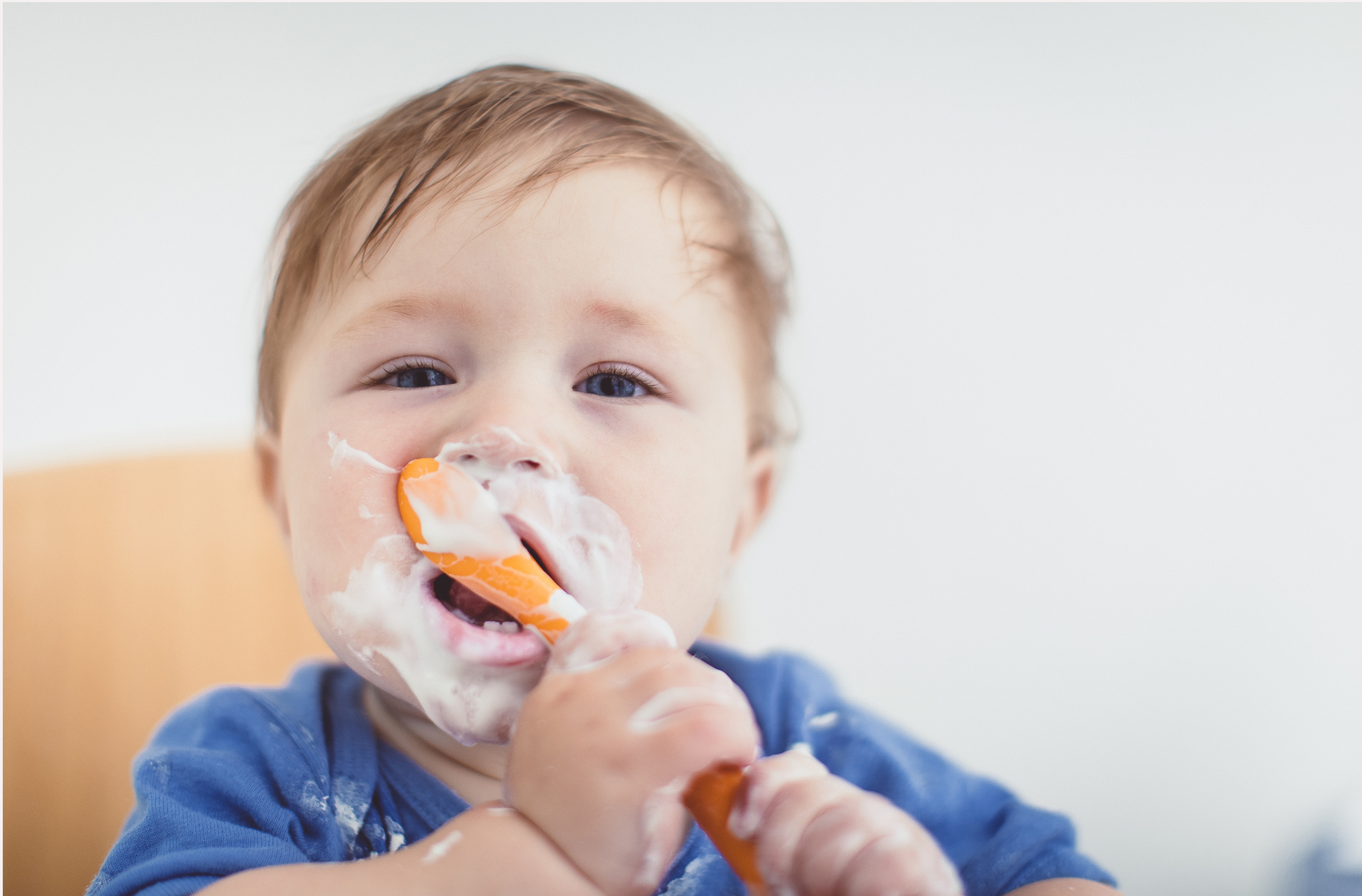 In the photo, a baby sits in a high chair, holding a spoon with one hand and enjoy eating yogurt. His face is adorned with yogurt, evident in the messy but joyful traces around his mouth and cheeks.